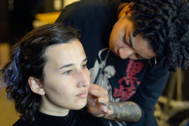 Afro makyöz kadın soyunma odasında genç bir mankene makyaj yapıyor.