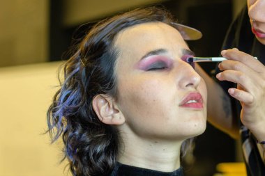 Genç bir kadın aynanın önünde bir akşam randevusu için makyaj yapıyor (kirpiklerini boyuyor). Onun yansımasına odaklan.