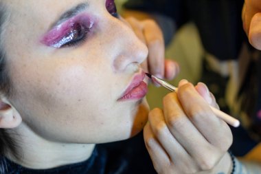 Genç bir kadın soyunma odasında profesyonel bir makyöz tarafından makyajlanıyor, dudaklarına ruj sürüyor.