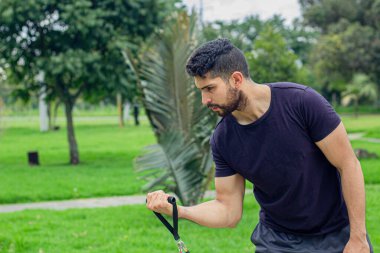 Lastik bantlarla antrenman yapan genç bir adam, parkta kol egzersizi yapıyor.