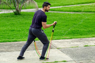 Lastik bantlarla antrenman yapan genç bir adam, parkta kol egzersizi yapıyor.