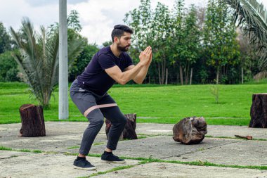 Lastik bantlarla antrenman yapan genç bir adam parkta bacak egzersizi yapıyor.