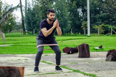 Lastik bantlarla antrenman yapan genç bir adam parkta bacak egzersizi yapıyor.