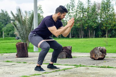 Lastik bantlarla antrenman yapan genç bir adam parkta bacak egzersizi yapıyor.