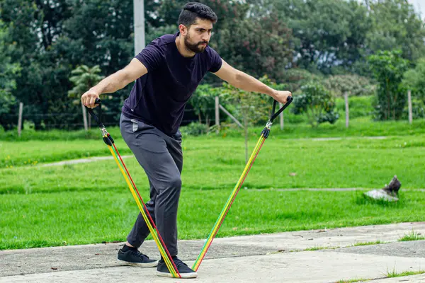 young man training with elastic bands, doing arm exercise outdoors in a park