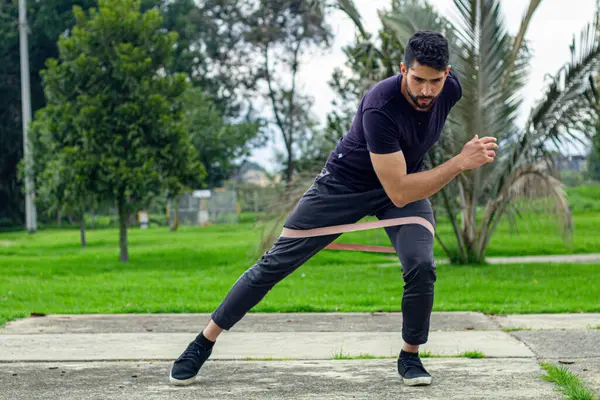young man training with elastic bands, doing squat leg exercise outdoors in a park