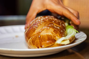 Fırıncının eli açık ekmekle sandviçi gösteriyor. Peynirli jambon, marul ve mayonezle kahvaltı masasında.