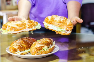 Fırıncının eli açık ekmekle sandviçi gösteriyor. Peynirli jambon, marul ve mayonezle kahvaltı masasında.