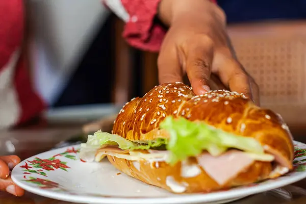 Fırıncının eli açık ekmekle sandviçi gösteriyor. Peynirli jambon, marul ve mayonezle kahvaltı masasında.