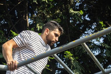 Parkta barda egzersiz yapan çekici bir genç adam kol egzersizi yapıyor.