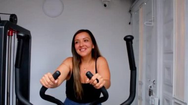 Spor salonunda eliptik bir makinede egzersiz yapan genç ve mutlu bir fitness kadını.
