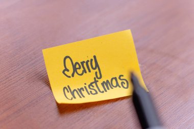 Bir insan masa tahtası arka planına yapışkan bir not yazıyor. Yazan kişi Mutlu Noeller diyor.