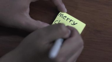 Bir insan masa tahtası arka planına yapışkan bir not yazıyor. Yazan kişi Mutlu Noeller diyor.