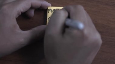 Bir insan masa tahtası arka planına yapışkan bir not yazıyor. Yazan kişi Mutlu Noeller diyor.