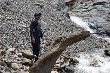 Sırt çantalı, siyah şapkalı bir adam dağ nehrinde bir şelalenin yanında duruyor ve yolculukta doğanın gücünün tadını çıkarıyor..