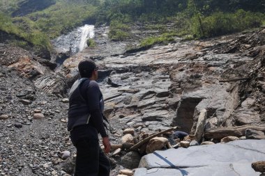 Sırt çantalı, siyah şapkalı bir adam dağ nehrinde bir şelalenin yanında duruyor ve yolculukta doğanın gücünün tadını çıkarıyor..