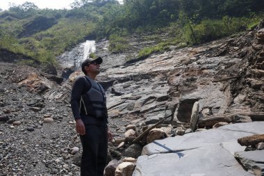 Sırt çantalı, siyah şapkalı bir adam dağ nehrinde bir şelalenin yanında duruyor ve yolculukta doğanın gücünün tadını çıkarıyor..