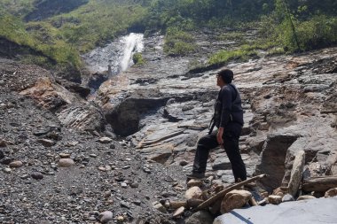 Sırt çantalı, siyah şapkalı bir adam dağ nehrinde bir şelalenin yanında duruyor ve yolculukta doğanın gücünün tadını çıkarıyor..