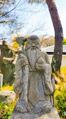 Kyoto, 25 Nisan 2025. Yaşlı sakallı bir adamın eskimiş taş heykeli. Elinde asa ve küre var. Güneşli bir bahçede..