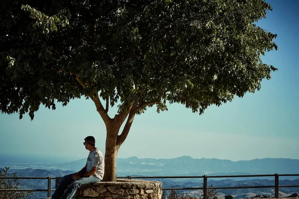 A Moment of Reflection: Man Sitting Under a Tree Admiring a Distant Mountainous Landscape