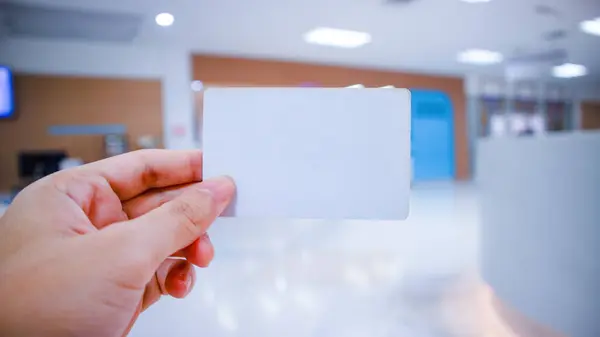 Female hand holding blank hospital card, business or discount card Mockup of hospital membership card, insurance card. Over the reception room of a hospital or clinic hospital, blurred in the bac
