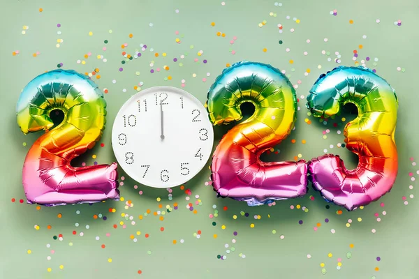 Colorful bright air balloon numbers and white watch at 12 oclock on light green background with confetti. 2023 New Year concept celebration