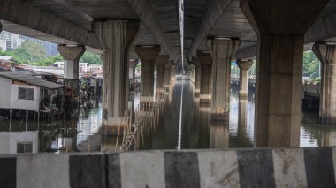 Büyük bir beton sütunun resmi, gözün görebildiği kadar yüksek iki rotayı destekler. Suyun altındaki alan büyük bir kanaldır. Bankaya tek katlı evler inşa eden köylüler var.