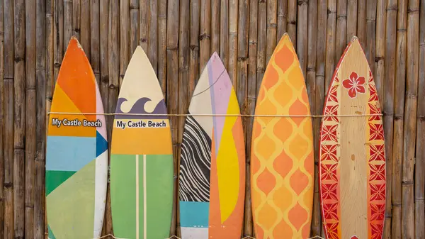A picture of five surfboards resting against a brown bamboo wall. Each surfboard has different colors, patterns, and sizes.