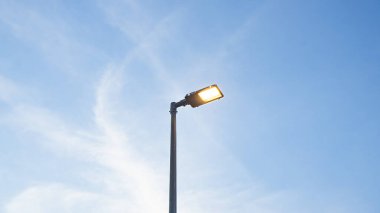 Yol kenarındaki uzun siyah ışık direğinin konum fotoğrafları. Resmin ortasında tek başına sıcak sarı bir ışık var. Arka plan, akşam mavisi gökyüzünün altında soluk bulutlardır. Atmosfer 