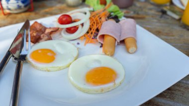 Otelin kahvaltı takımının resmi, kızarmış yumurta, pastırma, jambon ve sosis ile birlikte beyaz porselen tabakta. Yemeye hazır çatal ve bıçaklar var..