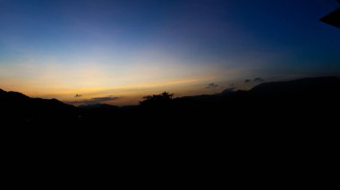 Sabah gökyüzü fotoğrafçılığı, gün doğumuyla mavi gökyüzü, altın sarısı ile çelişiyor. Gökyüzünde süzülen soluk bulutlar var. Uzaktaki siyah dağların ve büyük ağaçların gölgelerini görebilirsin.. 