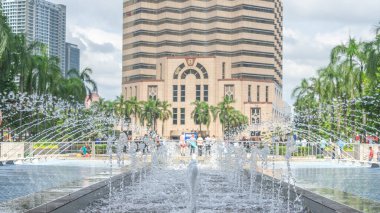 Yüksek bir binanın önündeki fıskiyede birçok çeşme var. Arka planda her yere su sıçrıyor. Arkadaki bina açık kahverengi, modern şekilli..