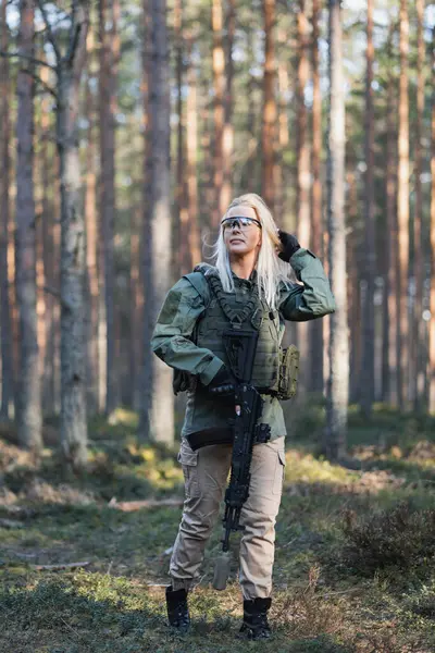 Beautiful blonde military girl with a modern AK12 suppressed rifle in the forest at war. Vertical photo.