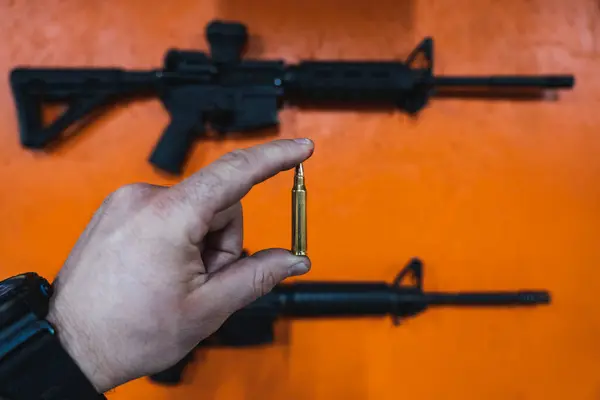 Shooting weapons in a shooting range. A 5.56x45mm cartridge in a man\'s hand and an m4a1 rifle on an orange wall.