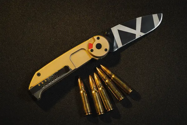 Tactical folding knife in camouflage color and rifle cartridges on a black background.