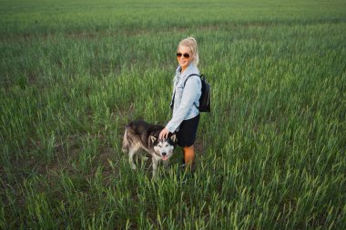 Güneş gözlüklü genç sarışın bir kız bir yaz akşamı bir köpekle birlikte tarlada yürüyor.. 