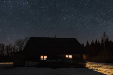 Silhouette of a forest wooden house with light in the windows at night with a starry sky.  clipart