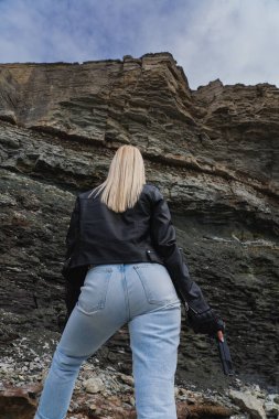 Deri ceketli, elinde silahla açık havada uçurumun kenarında bir kız. Arkadan görüntüle, dikey fotoğraf. 