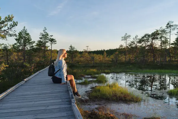 stock image A girl looks at the sunset while sitting on a wooden path in nature on the Viru swamp on a summer evening. 