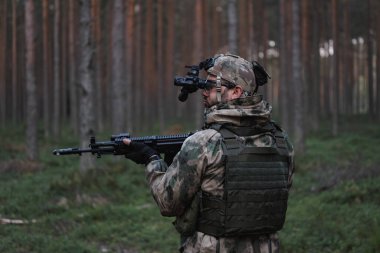 Kafasında gece görüşü cihazı ve alacakaranlıkta ormanda elinde AK-12 tüfeğiyle kamuflajlı ve çelik yelekli bir asker. Arka plan. 