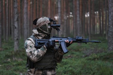 Kamuflajlı bir asker, kar maskesi ve çelik yelek, kafasında gece görüşü cihazı ve alacakaranlıkta ormanda elinde bir AK 12 tüfeğiyle..