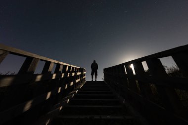 Gece sahnesi, kapüşonlu bir adamın silueti ay ışığı ve yıldızlı gökyüzünün altında doğada bir merdivende.