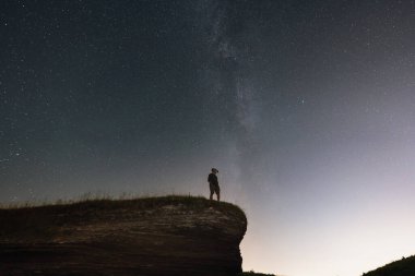 Gece sahnesi, şortlu bir adam ve gece görüş cihazı olan bir gömlek uçurumun kenarında duruyor. Yıldızlı gökyüzü ve Samanyolu. 