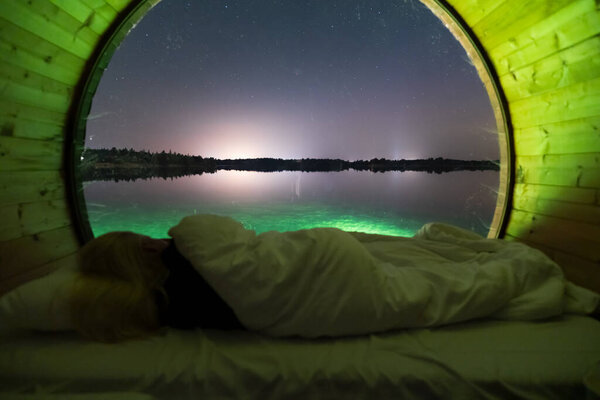 Night photo, girl sleeping on a bed in a barrel house on the water, large window overlooking the lake and the starry sky.