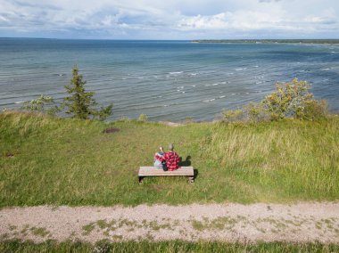 A young couple sits on a bench on the steep shore of the Baltic Sea on a summer day, photo view from a drone.  clipart