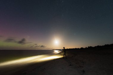 El feneri olan adam kumlu sahilde duruyor yıldızlı gökyüzü ve ay, manzara astrofotografı.