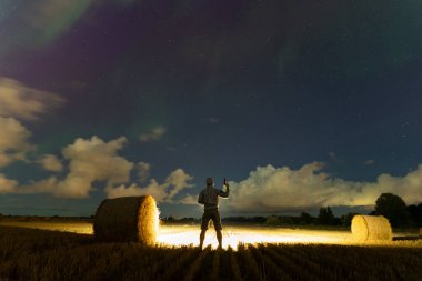 Night scene, a man with a gun in his hand in a field with haystacks under a starry sky with northern lights. View from behind.  clipart