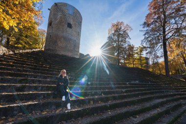 A young girl of European appearance sits on a stone staircase near the Cesis castle on an autumn morning. Bright sunbeams shine into the camera lens.  clipart