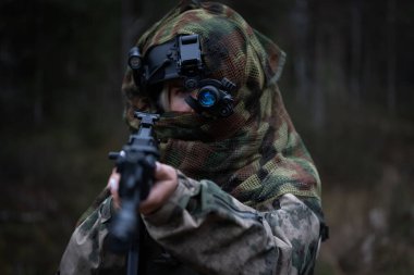 Kamuflajlı bir kadın asker, üzerinde gece görüşü cihazı olan bir kask ve elinde bir saldırı tüfeği olan karanlık bir ormanda..