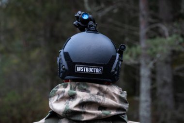 Military instructor in a tactical helmet with a night vision monocular in the forest at dusk. View from the back. clipart